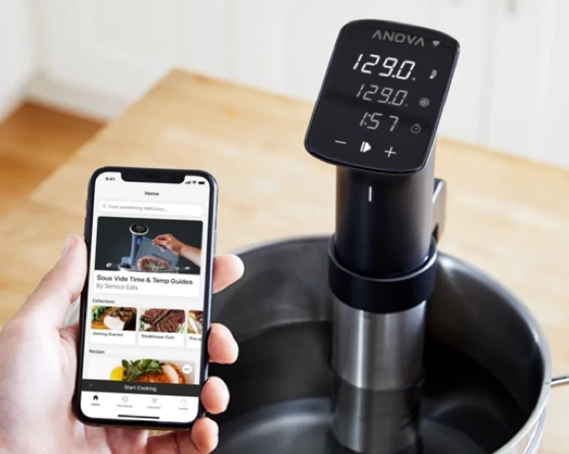 Anova Precision Cooker Pro set up in a pot of water, with a hand holding a smartphone displaying the Anova app for remote sous vide control.